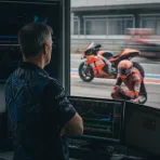 A senior MotoGP team manager seen in his pit box office, with a rival's red motorcycle visible in the background.