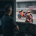 A senior MotoGP team manager seen in his pit box office, with a rival's red motorcycle visible in the background.