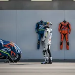 A MotoGP rider in a pit garage contemplates a choice between a blue Yamaha racing suit and an orange Honda one.