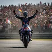 A MotoGP rider celebrates a Grand Prix victory in 2026, standing on his motorcycle in front of a cheering crowd.