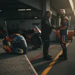 A rider's helmet sits on a pit wall, with the out-of-focus shape of a person in the garage behind, symbolising mentorship in MotoGP.