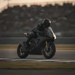 A MotoGP-style racing bike leans low through a corner at sunset, creating sparks as the rider's knee touches the track.