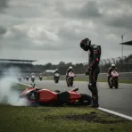 A dejected MotoGP rider stands beside his broken-down race bike at the side of the track.