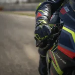 A close-up photograph of a MotoGP rider's gloved hand under strain as it grips the handlebar of their racing motorcycle.