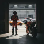 A rider in racing leathers stands in a pit garage, looking at a brand new MotoGP motorcycle ready for the 2027 season.