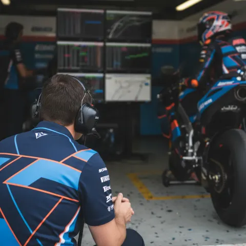 A MotoGP team manager watches timing screens with intense focus from inside the pit box during a race weekend.