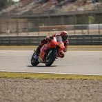 A MotoGP rider leans forward over the handlebars of their red racing bike at high speed on a circuit's main straight.